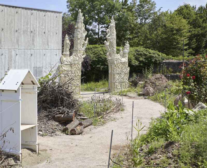 Jardin du FRAC, un chemin de terre se dirige vers un portrait en céramique constitué de formes abstraites et orientales. Un rosier, et une maisonnette blanche habille le jardin.