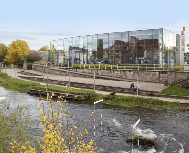 Extérieur du FRAC Alsace, c'est l'été, une rivière passe devant le bâtiment. La façade est composé de fenêtres, la ville se reflète dedans.