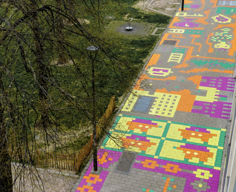 Rue vue de haut : les pavés ont été coloriés en différentes couleurs sur qui crée des dessins vue du dessus : des fleurs, des personnages, un serpent, des formes abstraites.