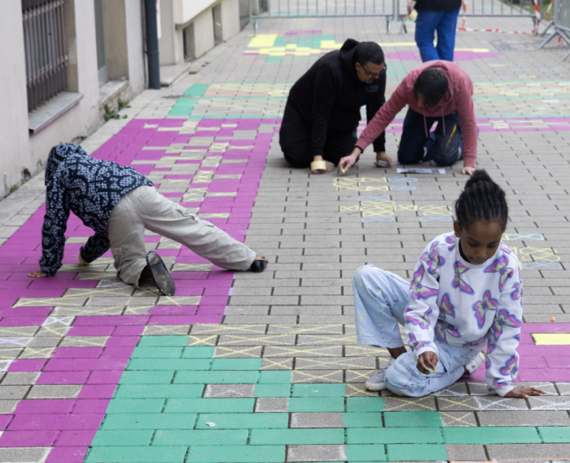 Sur une route pavée, un monsieur, et 3 enfants dessinent sur le sol à la craie. Les couleurs sont pétillantes : fuchsia, turquoise et jaune.