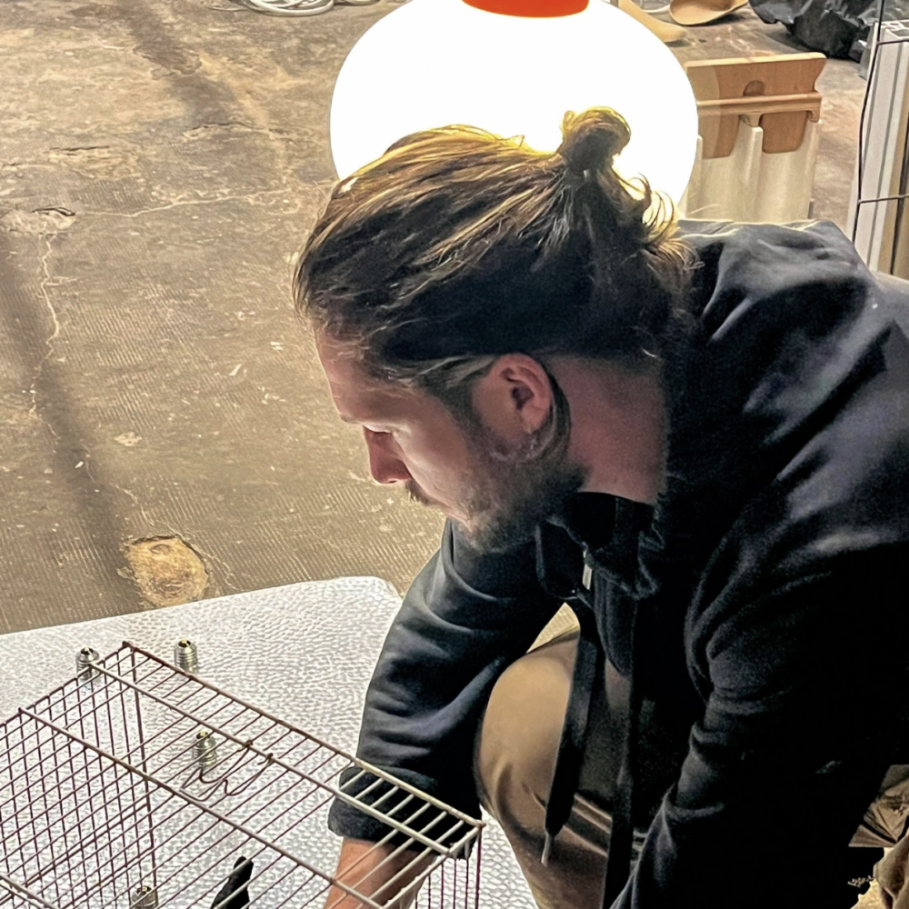 Vincent Gallais est de profil et travaille sur une sculpture : il est concentré et il installe une cage en fer sur un support en acier.