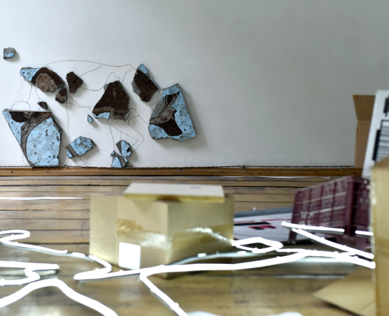 Installation de Vincent Gallais : au sol des néons dessinent une forme abstraite, par dessus : des cartons, des papiers avec des illustrations. Au loin contre le mur, des fils de fer relient des blocs de carrelage bleu clair.