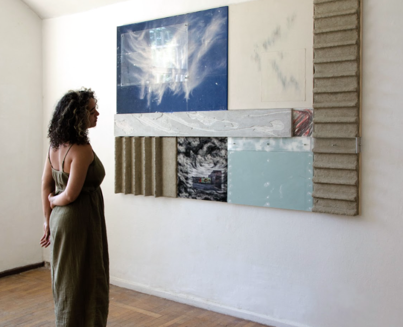 Une femme contemple un tableau de Vincent Gallais. Le tableau est composé de plusieurs rectangles. Chaque rectangle a un matière différente et un aspect différent : un ciel bleu, du carton, du béton, une couleur unie.