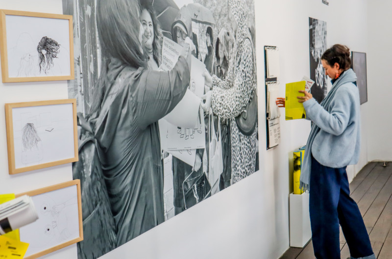 Une femme visite une exposition. Sur le mur une grande photo en noir et blanc représentante des femmes militantes. Autour de la photo, des illustrations.