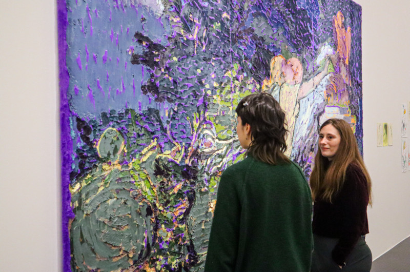 Deux femmes sous devant un grand tableau. Le tableau est un tapis géant violet sur lequel l'artiste à peint une scène où une femme et un homme s'embrasse dans un jardin luxuriant.